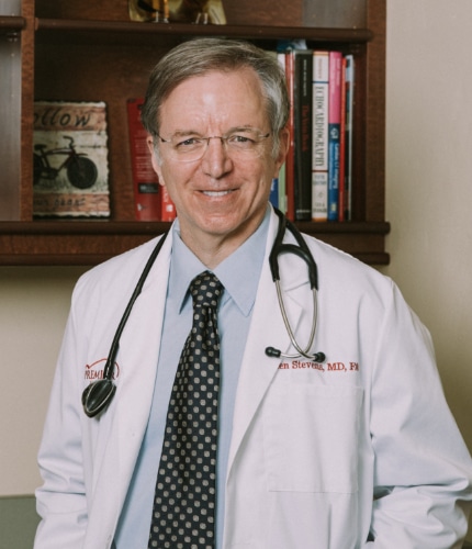 doctor standing with white coat on smiling with bookshelf in background