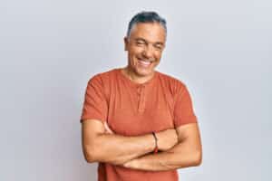 A middle aged man smiles and poses for the camera with his arms crossed against a gray background