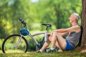Senior cyclist sitting by a tree in a park with a water bottle in his hand